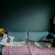 Worn blue couch in dim room with a pink blanket, pillows, and open laptop, like someone has been sitting there recently.